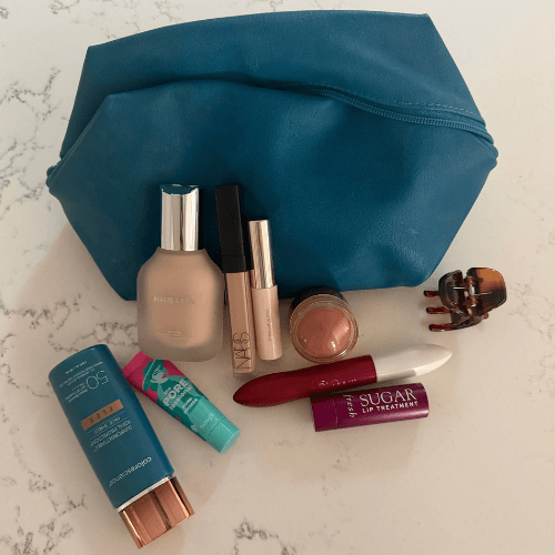 5-minute makeup routine products in a teal makeup bag on a marble counter: tinted SPF, primer, foundation, concealer, brow gel, blush, mascara, and lip balm.